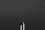 Two writing instruments, a pencil and a pen, are positioned next to each other on a textured dark surface, with ample space surrounding them.