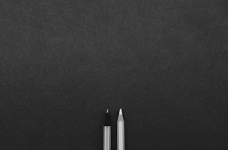 Two writing instruments, a pencil and a pen, are positioned next to each other on a textured dark surface, with ample space surrounding them.