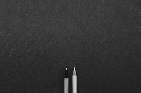 Two writing instruments, a pencil and a pen, are positioned next to each other on a textured dark surface, with ample space surrounding them.