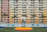 A colorful basketball court is set against the backdrop of a residential building featuring a grid of multicolored balconies. Several palm trees line the court, adding a touch of greenery to the urban scene.