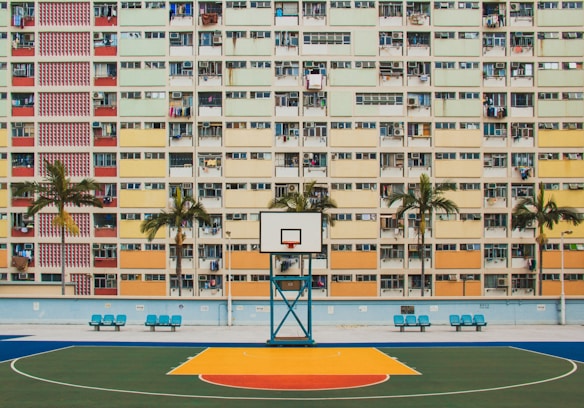 A colorful basketball court is set against the backdrop of a residential building featuring a grid of multicolored balconies. Several palm trees line the court, adding a touch of greenery to the urban scene.
