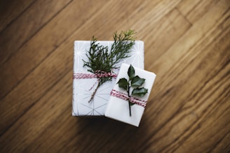 Two wrapped gifts with natural decorations are placed on a wooden floor. The larger gift is adorned with a sprig of evergreen and red and white twine, while the smaller gift has a holly leaf attached with similar twine. The wrapping paper is a minimalist geometric design.