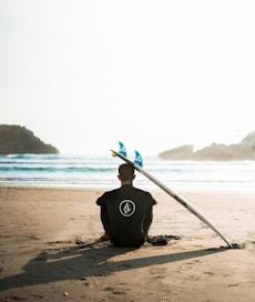 man sitting on sand beside surfboard facing sea during daytime