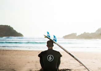man sitting on sand beside surfboard facing sea during daytime