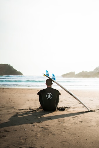 man sitting on sand beside surfboard facing sea during daytime