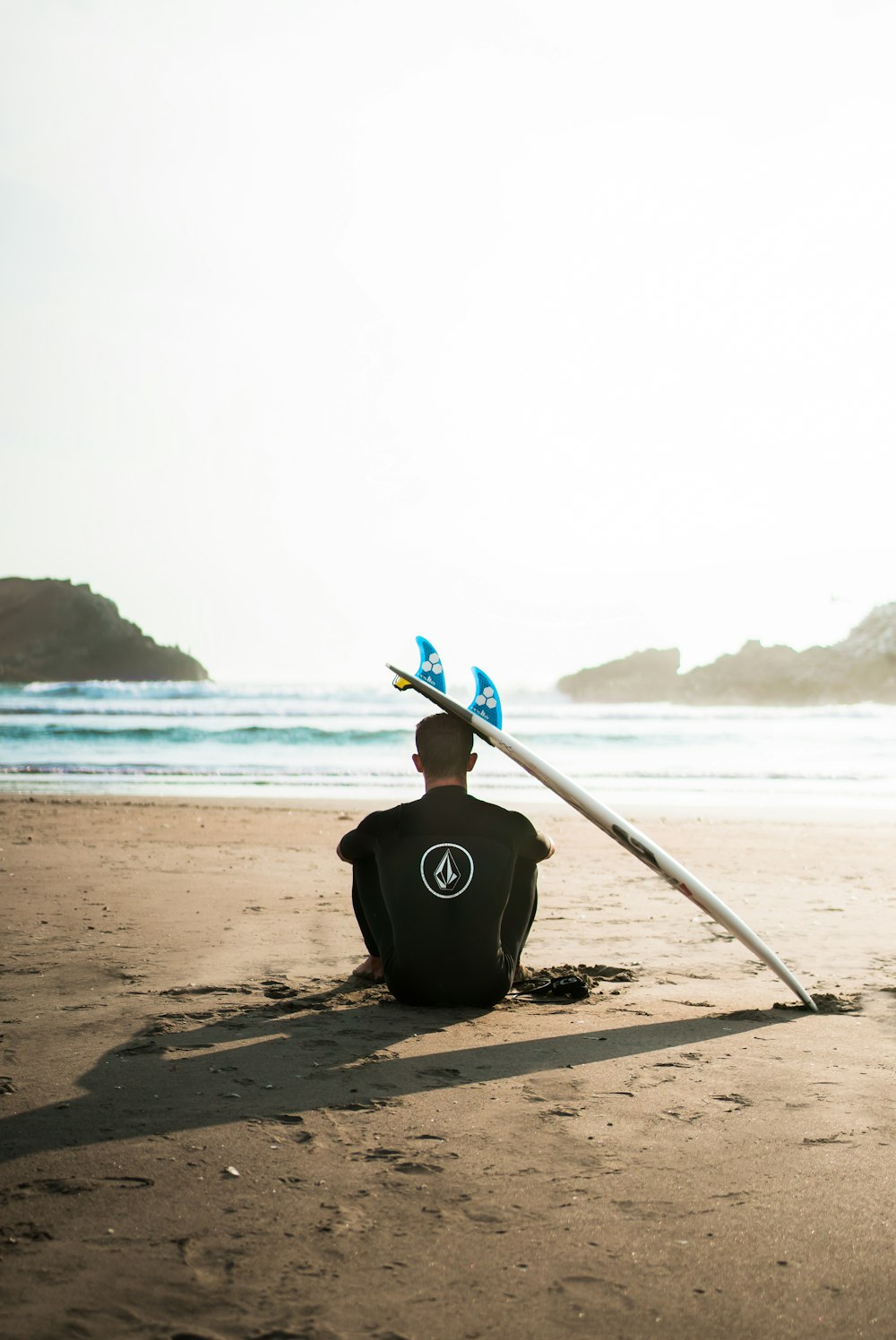 man sitting on sand beside surfboard facing sea during daytime