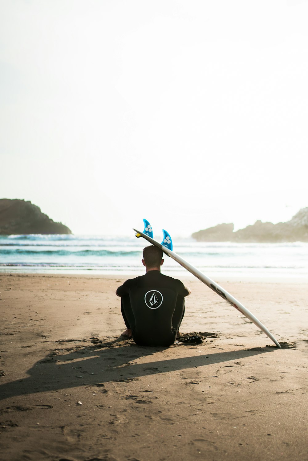 homem sentado na areia ao lado da prancha de surf de frente para o mar durante o dia
