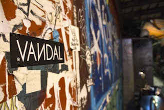 A close-up view of a wall covered in peeling posters, graffiti, and stickers, with a prominent black and white sticker labeled 'VANDAL' in bold letters at the forefront. The background is blurred but shows more graffiti in vibrant colors, creating a chaotic and urban atmosphere.