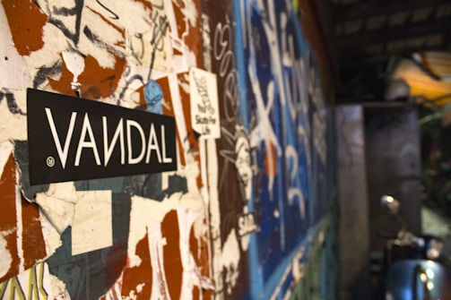 A close-up view of a wall covered in peeling posters, graffiti, and stickers, with a prominent black and white sticker labeled 'VANDAL' in bold letters at the forefront. The background is blurred but shows more graffiti in vibrant colors, creating a chaotic and urban atmosphere.