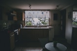 A dimly lit kitchen with wooden cabinets and an L-shaped countertop featuring a sink in front of a large window. The window has a patterned curtain and offers a view of trees outside. A small table with a plant on top is visible in the foreground.