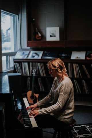 A person with blonde hair wearing glasses plays the piano in a cozy room. The wall behind features a collection of vinyl records, a guitar leaning against the wall, and framed art. Soft lighting creates a warm, intimate atmosphere.