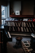 A cozy music room with a collection of vinyl records neatly organized on shelves. A mandolin hangs on the wall above the records, accompanied by a framed artwork. A piano is partially visible on the left side, and a vintage stool is centrally placed on a patterned rug.