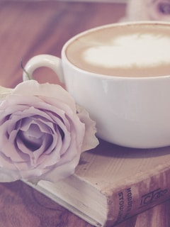 A cup of cappuccino with a frothy top sits on a closed book, accompanied by a delicate light pink rose. The scene is serene and softly lit, with a warm wooden background adding to the cozy ambiance.
