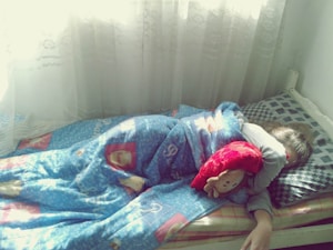 A person is sleeping on a bed with a blue patterned blanket and a pillow under their head. They are holding a red plush toy, and the room is softly lit by natural light coming through semi-transparent curtains.