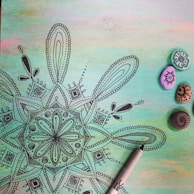 A detailed black mandala drawing on a pastel-colored background with shades of green, blue, and pink. To the right, there are four small painted stones with intricate designs in black ink. A black marker pen is partially visible at the bottom right corner.