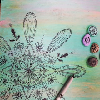 A detailed black mandala drawing on a pastel-colored background with shades of green, blue, and pink. To the right, there are four small painted stones with intricate designs in black ink. A black marker pen is partially visible at the bottom right corner.
