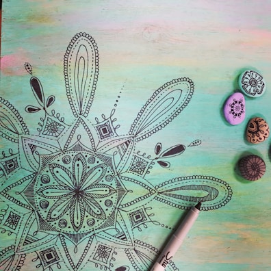 A detailed black mandala drawing on a pastel-colored background with shades of green, blue, and pink. To the right, there are four small painted stones with intricate designs in black ink. A black marker pen is partially visible at the bottom right corner.