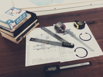 A wooden desk is covered with technical drawings featuring mechanical parts. Several measuring tools, including a digital caliper, are placed on top of the papers alongside a roll of tape and a tape measure. A stack of books with Russian titles sits at one corner of the desk.