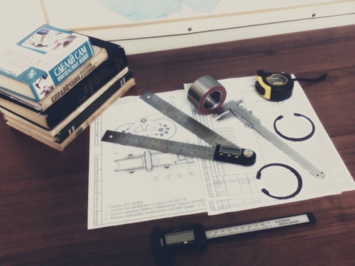 A wooden desk is covered with technical drawings featuring mechanical parts. Several measuring tools, including a digital caliper, are placed on top of the papers alongside a roll of tape and a tape measure. A stack of books with Russian titles sits at one corner of the desk.