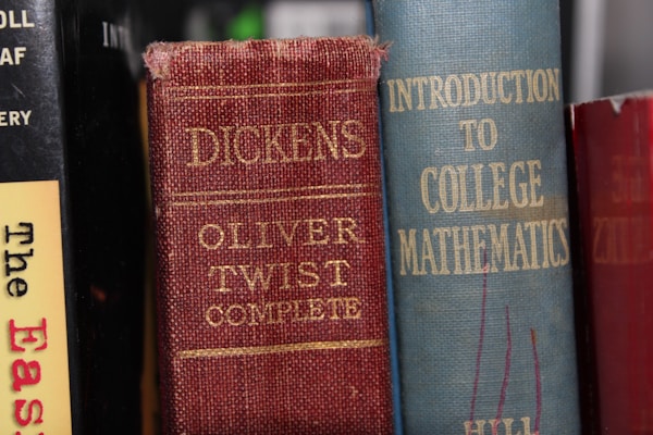 Several books are aligned vertically on a shelf. The visible titles include 'Oliver Twist Complete' by Dickens with a red and worn cover, alongside 'Introduction to College Mathematics' which has a blue cover. Another book with a partially visible title, 'The Eas...', has a yellow and black cover.