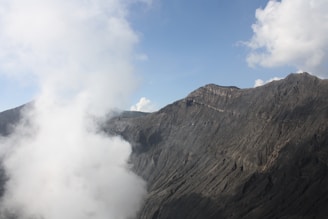 A rugged mountain terrain with steep, textured slopes against a clear blue sky. Billowing clouds of white smoke or mist rise prominently in the foreground.