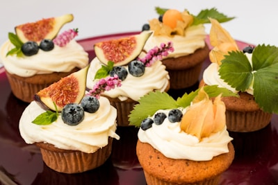 A group of six cupcakes, each topped with creamy white frosting and garnished with various fresh fruits and leaves. The toppings include sliced figs, blueberries, and decorative herbs. Some cupcakes also feature physalis with its distinctive husk. The plate is a deep red, adding a rich background to the vibrant colors of the desserts.