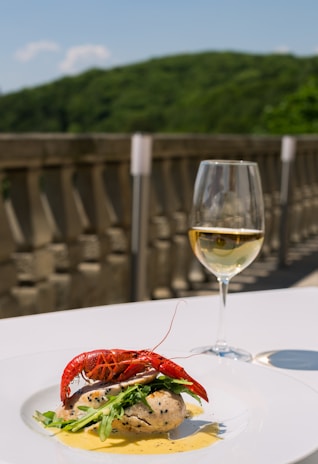A gourmet dish featuring a lobster on top of seasoned fish and greens, elegantly plated with a creamy sauce. A glass of white wine accompanies the meal, set against an outdoor backdrop with blurred foliage and a stone railing.