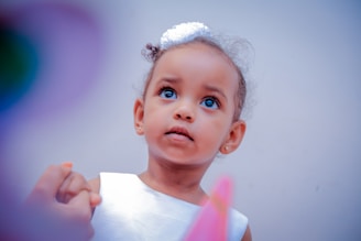 A young child with curly hair adorned with a small white accessory looks upwards with a thoughtful expression. She is wearing a white dress and is holding someone's hand, suggesting a tender moment. The background is a soft, blurred gradient that enhances the focus on the child.
