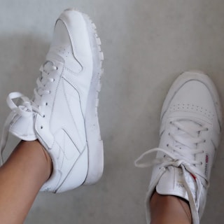 A pair of white sneakers with minimal design and perforated detailing is worn by a person. One shoe is positioned upright while the other is seen from the side, showing laces and a clean finish.