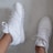 A pair of white sneakers with minimal design and perforated detailing is worn by a person. One shoe is positioned upright while the other is seen from the side, showing laces and a clean finish.
