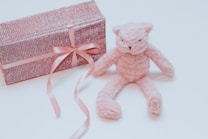 A soft, plush pink teddy bear is placed next to a rectangular, pink glittery gift box wrapped with a pink ribbon and bow. The items are set against a light, neutral background, creating a gentle and serene atmosphere.