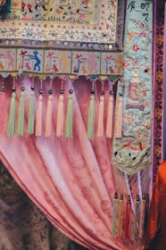 An intricately designed textile featuring colorful embroidery and patterns. The fabric showcases detailed motifs, including depictions of human figures, floral designs, and animals, bordered by decorative tassels in shades of pink, green, and yellow. The background is a pink draped curtain with subtle floral patterns.