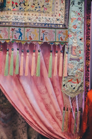 An intricately designed textile featuring colorful embroidery and patterns. The fabric showcases detailed motifs, including depictions of human figures, floral designs, and animals, bordered by decorative tassels in shades of pink, green, and yellow. The background is a pink draped curtain with subtle floral patterns.