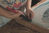 A person is using a tool with a red handle, possibly a staple remover or similar tool, to work on a wooden frame. The person is wearing a shirt with a colorful floral pattern, and their focus appears to be on the task at hand. The setting suggests a crafts or DIY environment.