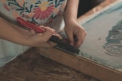 A person is using a tool with a red handle, possibly a staple remover or similar tool, to work on a wooden frame. The person is wearing a shirt with a colorful floral pattern, and their focus appears to be on the task at hand. The setting suggests a crafts or DIY environment.