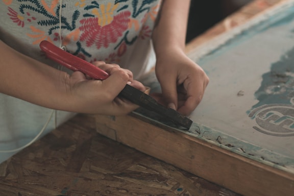 A person is using a tool with a red handle, possibly a staple remover or similar tool, to work on a wooden frame. The person is wearing a shirt with a colorful floral pattern, and their focus appears to be on the task at hand. The setting suggests a crafts or DIY environment.
