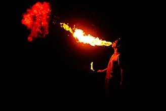 A person is performing a fire-breathing act, expelling a large burst of bright orange and red flames from their mouth. The surroundings are dark, highlighting the vivid colors of the fire.
