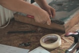 A person is using pliers on a wooden surface, with various tools and items nearby including a roll of tape, a pen, and memory cards. A piece of paper with handwritten notes is also visible.