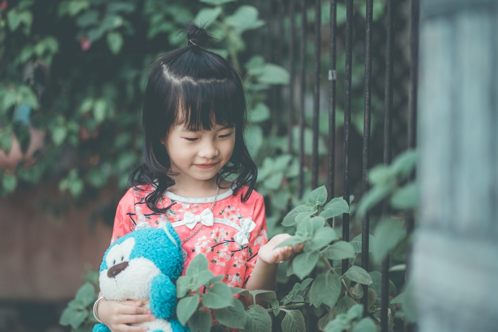 toddler girl holding leaf and plush toy near fence during daytime