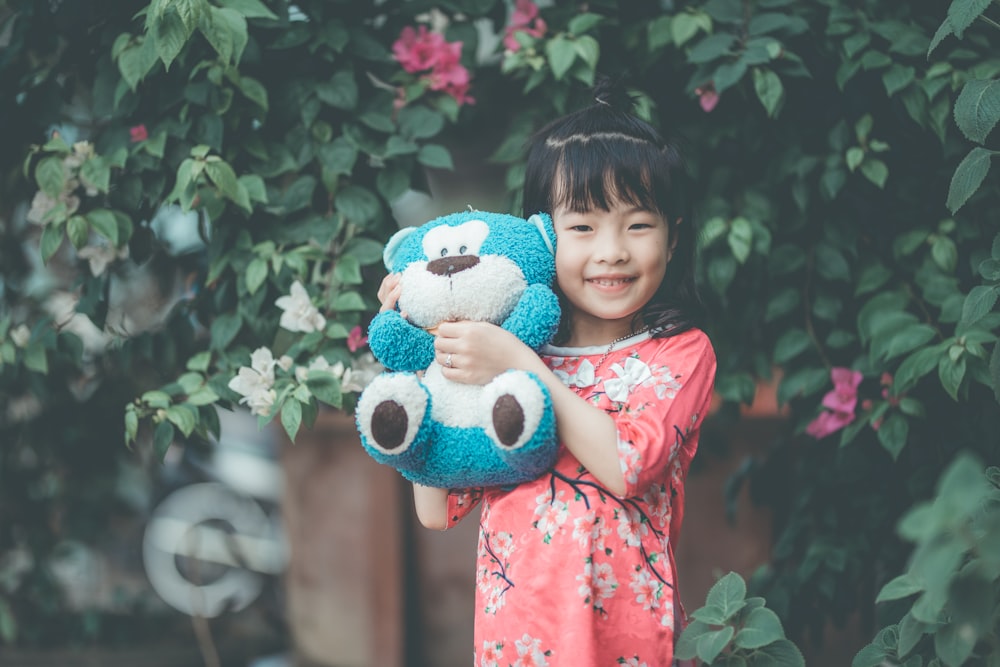 niña sosteniendo un juguete de peluche azul y blanco mientras está de pie cerca de una planta verde durante el día