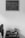 A black and white photograph featuring a small wooden table against a plain wall. On the wall hangs a chalkboard with the words "Toda pregação exige uma resposta" attributed to Pr. Luiz. On the table, there is a stack of books, a cup holding paintbrushes, and a small decorative object. To the left, an office chair is partially visible.
