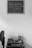 A black and white photograph featuring a small wooden table against a plain wall. On the wall hangs a chalkboard with the words "Toda pregação exige uma resposta" attributed to Pr. Luiz. On the table, there is a stack of books, a cup holding paintbrushes, and a small decorative object. To the left, an office chair is partially visible.