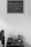 A black and white photograph featuring a small wooden table against a plain wall. On the wall hangs a chalkboard with the words "Toda pregação exige uma resposta" attributed to Pr. Luiz. On the table, there is a stack of books, a cup holding paintbrushes, and a small decorative object. To the left, an office chair is partially visible.