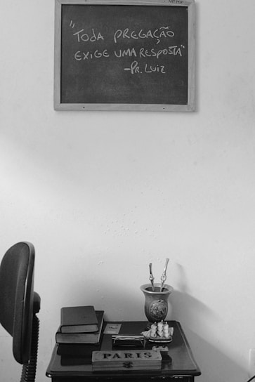 A black and white photograph featuring a small wooden table against a plain wall. On the wall hangs a chalkboard with the words "Toda pregação exige uma resposta" attributed to Pr. Luiz. On the table, there is a stack of books, a cup holding paintbrushes, and a small decorative object. To the left, an office chair is partially visible.