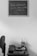 A black and white photograph featuring a small wooden table against a plain wall. On the wall hangs a chalkboard with the words "Toda pregação exige uma resposta" attributed to Pr. Luiz. On the table, there is a stack of books, a cup holding paintbrushes, and a small decorative object. To the left, an office chair is partially visible.