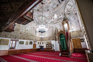 An ornate interior space with intricate floral and geometric patterns covering the walls and ceiling. The space features a richly decorated wooden pulpit, adorned with a green cloth, and a chandelier hanging from the center. The floor is covered with a red patterned carpet. Natural light filters through small windows, and framed calligraphy art hangs on the walls.