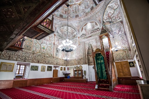 An ornate interior space with intricate floral and geometric patterns covering the walls and ceiling. The space features a richly decorated wooden pulpit, adorned with a green cloth, and a chandelier hanging from the center. The floor is covered with a red patterned carpet. Natural light filters through small windows, and framed calligraphy art hangs on the walls.