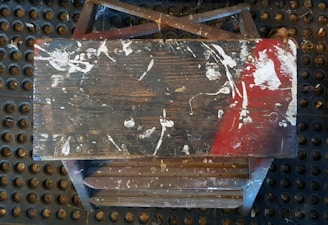 A worn-out, paint-speckled wooden bench seat is placed atop a rubber mat with circular patterns. The bench features prominent red and white paint stains, with visible scratches and markings across its surface.