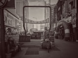 A black and white urban scene featuring tall buildings, a street with various signs in Chinese, and a large archway with promotional banners. Several pieces of furniture and a cart are placed in the foreground, along with potted plants and a variety of small shops on the side.