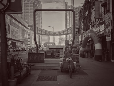 A black and white urban scene featuring tall buildings, a street with various signs in Chinese, and a large archway with promotional banners. Several pieces of furniture and a cart are placed in the foreground, along with potted plants and a variety of small shops on the side.